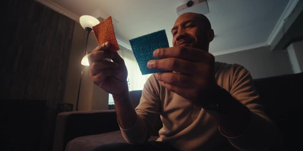 A man stands in a room holding two rectangular objects likely fabric samples as he examines and compares them