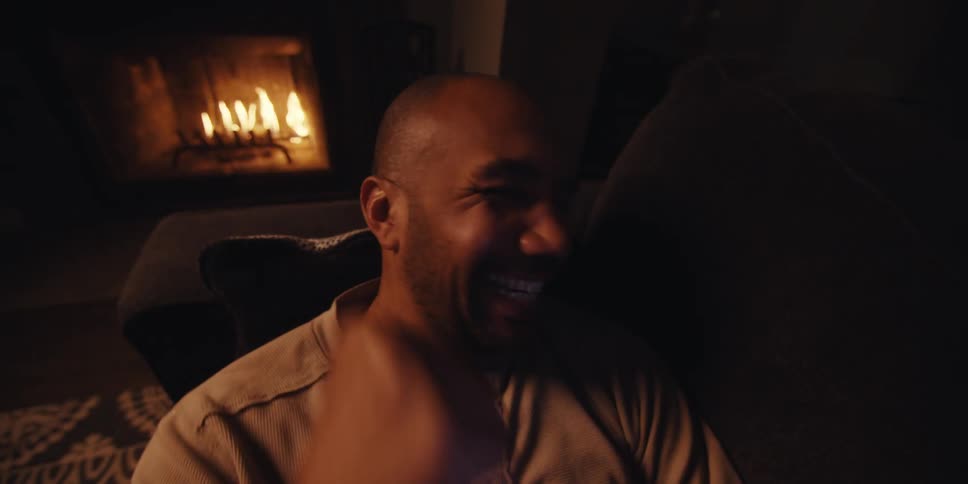 Man lying down on a couch near a lit fireplace celebrates while watching video on his smartphone