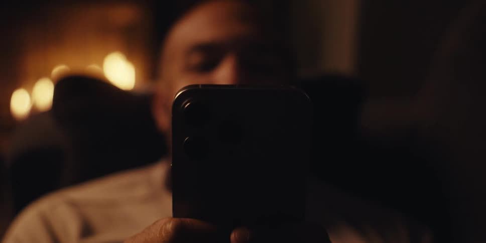 Man lying on his couch views his smartphone screen while a fire burns in the fireplace