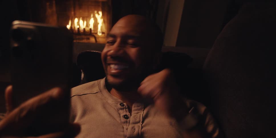 A man sits on a couch in a cozy room with a fireplace holding a phone and celebrating joyfully