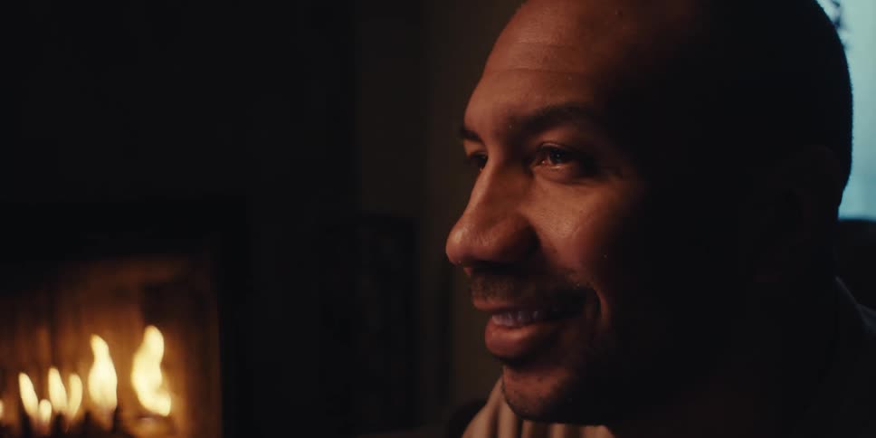 The face of a man who is giving a smile looking forward and behind him there is a fireplace