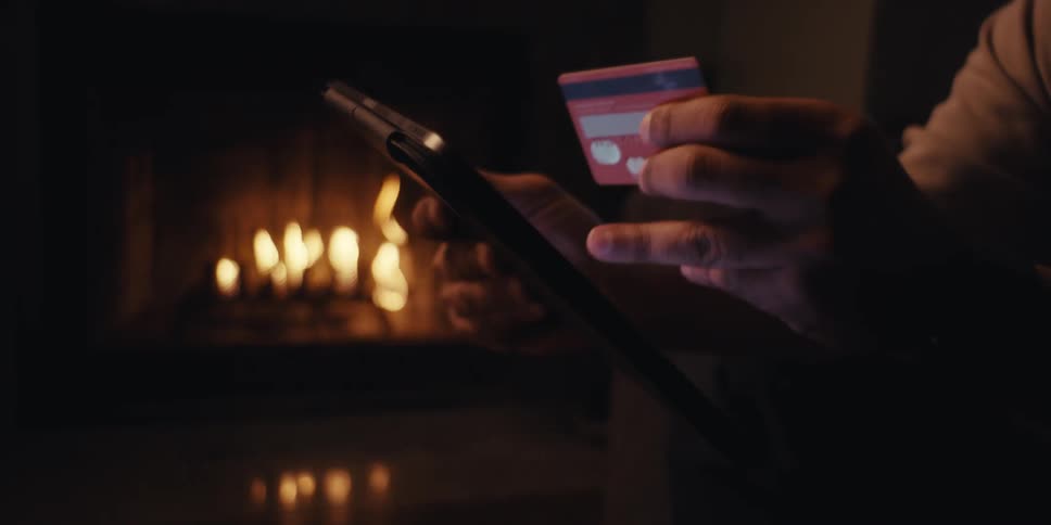 A man sits near a fireplace in a dimly lit room making a payment on a tablet with his credit card