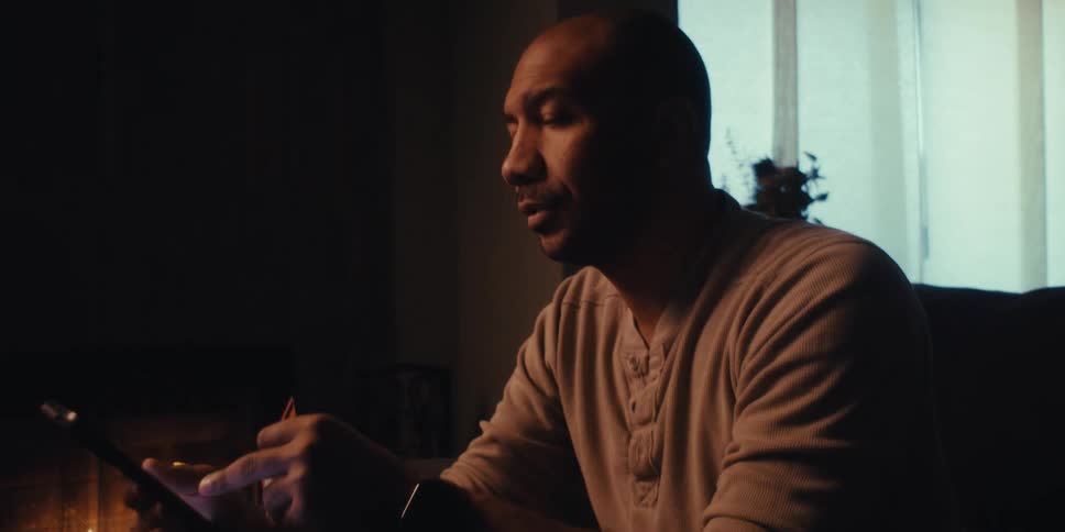 A man using a cell phone while sitting near a hill in a partially dark room with a window behind