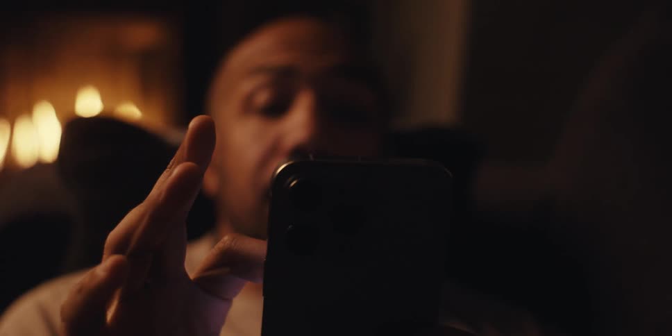 A man sits on a couch in a cozy room with a fireplace holding a phone