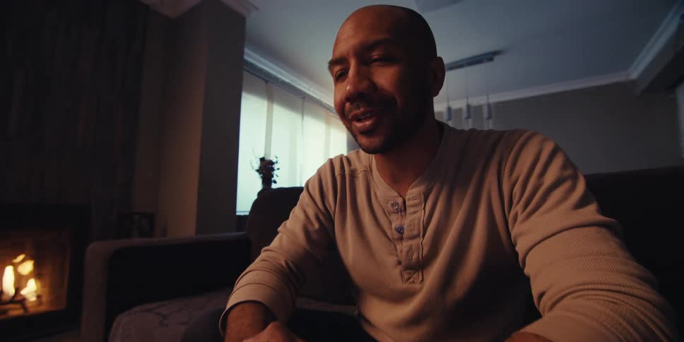 A man with a shaved head short beard and light colored sweatshirt sits in a dimly lit room illuminated by the warm glow of a fireplace having a video call on a tablet on the coffee table