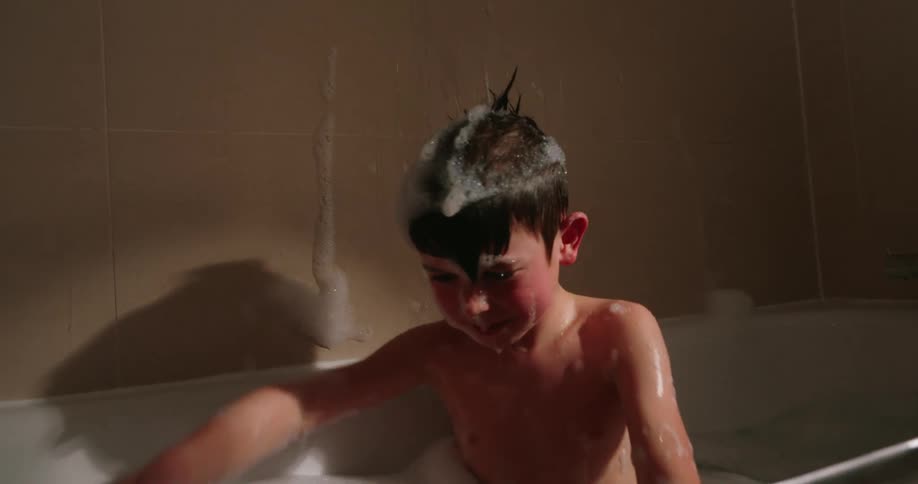 A boy splashes water and foam playfully with his hands while taking a bath in a bathtub