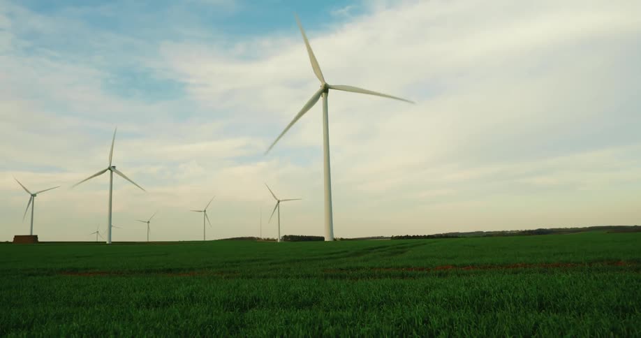 From a low vantage in a farmland field the view sweeps upward to reveal tall wind turbines spinning against a broad sky their sleek blades punctuating the open landscape as they harness the breeze