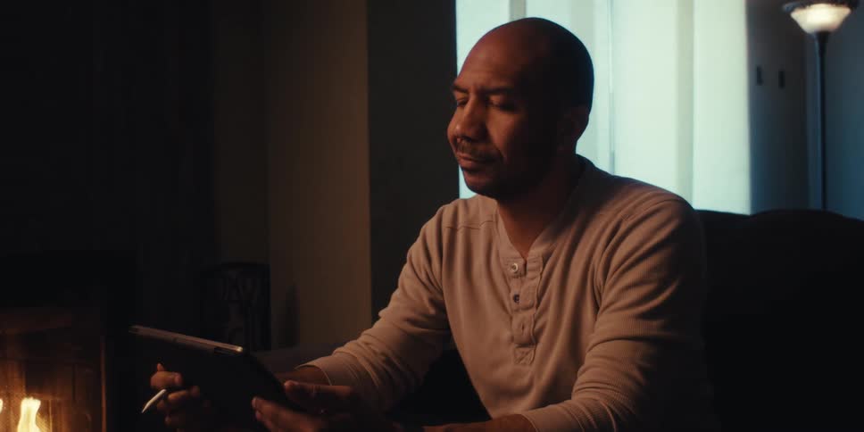 A man sits on a couch in a dimly lit room near a fireplace intently focused on a drawing or writing tablet in his hands