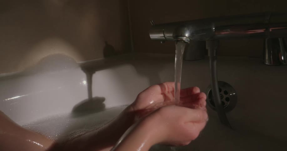 A boy sits in a dimly lit bathtub using his hands to wash his face with water flowing from the faucet
