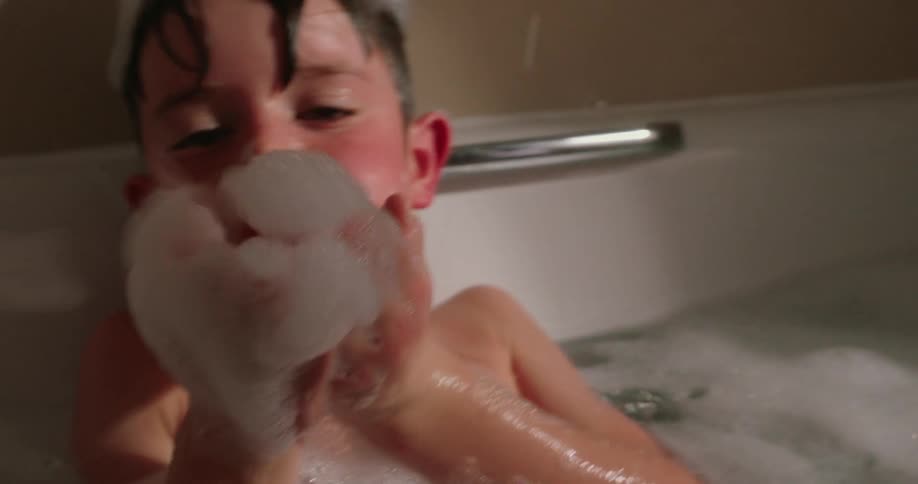 Boy blows the bubbles held in his hands while taking a bubble bath