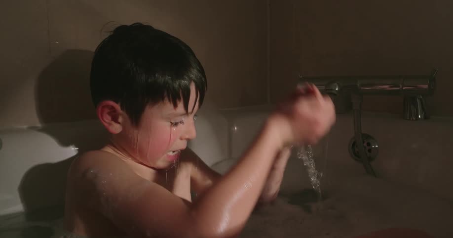A boy in a bathtub splashes water over his head with his hands as it flows from a faucet