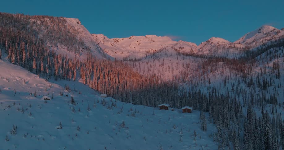A serene snow covered mountain landscape at dusk features a small cluster of cabins nestled among snow laden trees with the peaks catching the last light of the setting sun