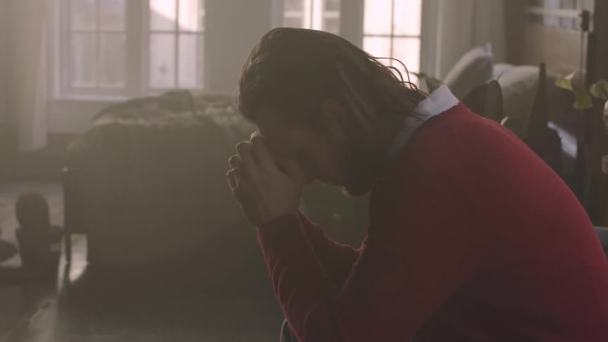 A person clasps their hands together possibly in a gesture of prayer or contemplation with a ring visible on one finger set against a softly lit background
