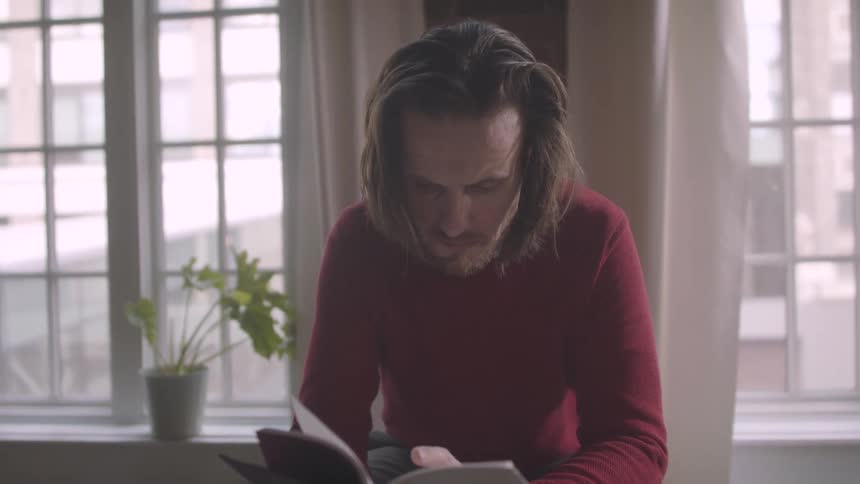 A man with long hair reads a book in a room with a window and a potted plant