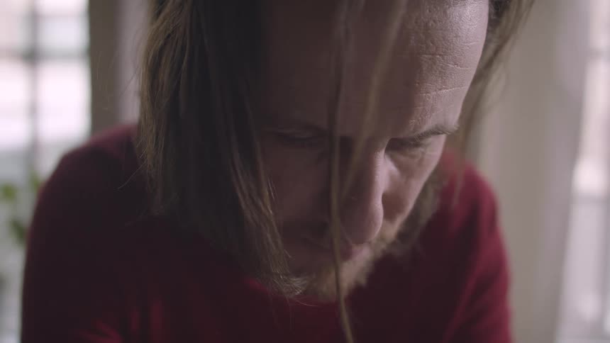 A middle aged man with long hair a beard and a red sweater looks down in the room