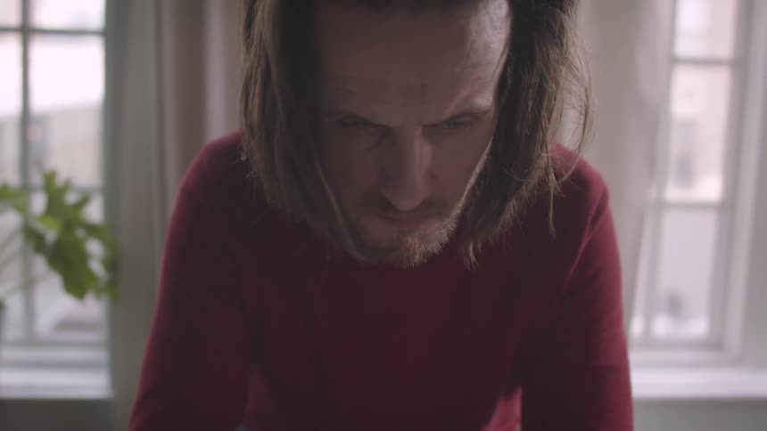 A bearded man with long hair stands still in front of windows providing a softly lit background