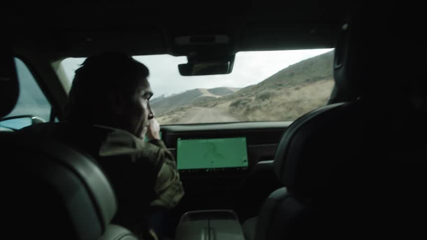 A person is driving a vehicle through a mountainous landscape with a large touchscreen display showing a map on the dashboard