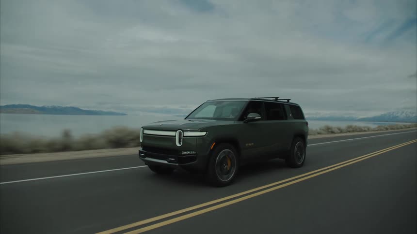 A dark colored rivian r1s suv drives along an asphalt road next to a large body of water with mountains in the distance under an overcast sky