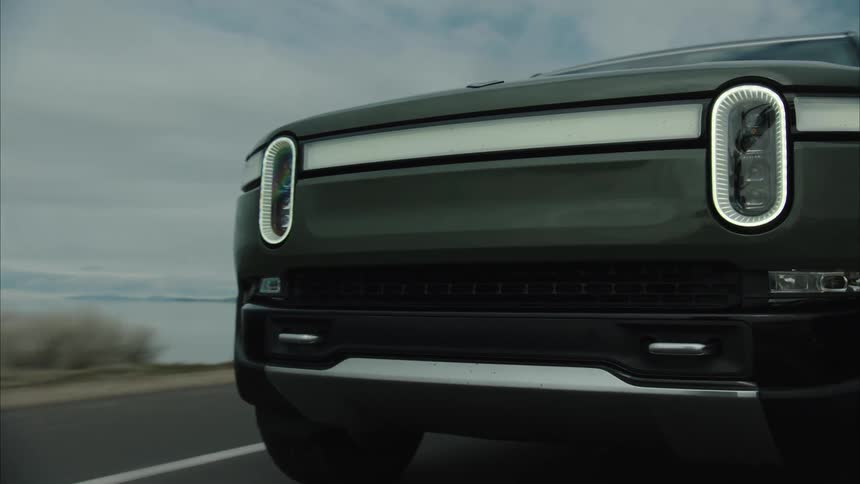 A dark rivian r1s travels along a paved road through a vast lakeside landscape beneath an overcast sky