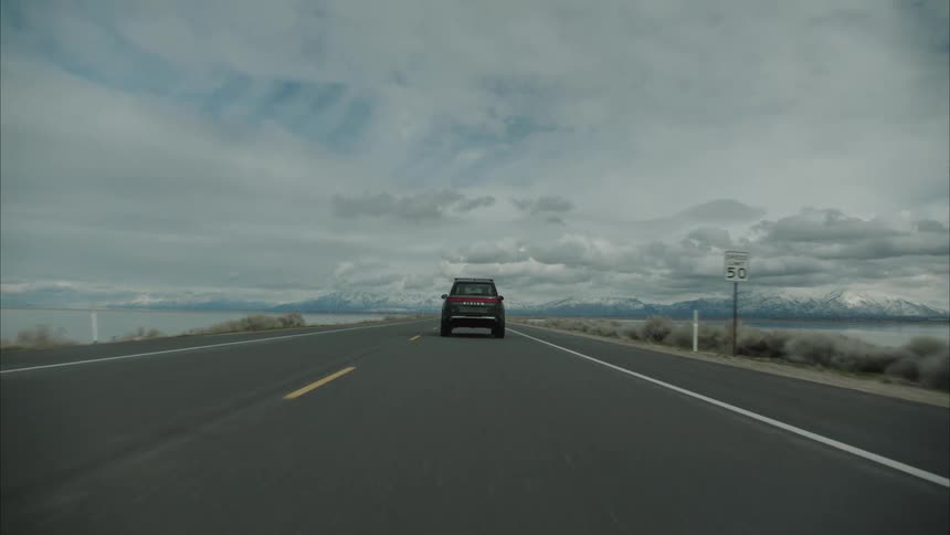 A dark rivian r1s drives along an asphalt road next to a large body of water with mountains in the distance under an overcast sky