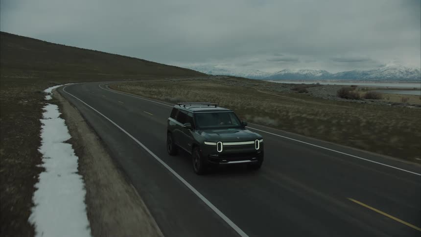 A dark colored rivian r1s suv drives along a winding asphalt road through a barren landscape towards a lake with snowcapped mountains in the distance under a cloudy sky