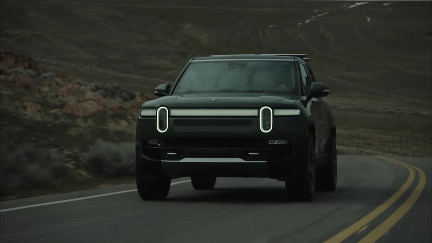 A dark colored rivian r1s suv drives along an asphalt road through a dry hilly landscape