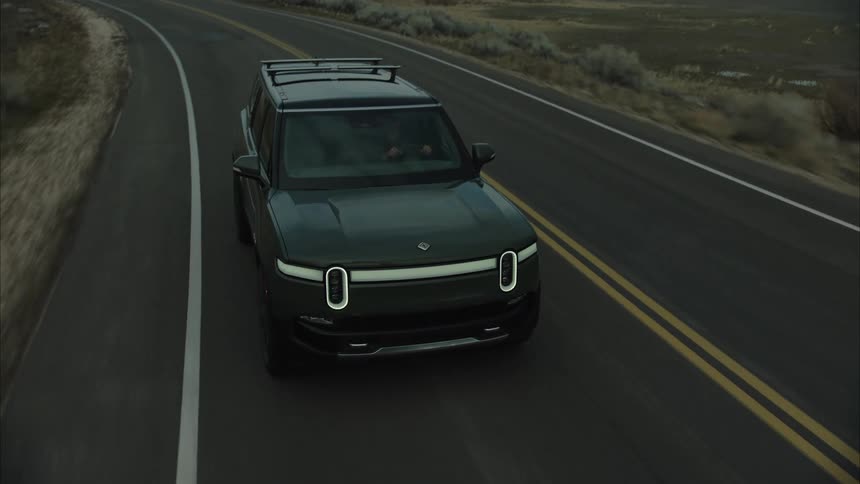 A dark rivian r1s suv drives along a straight asphalt road through a barren landscape with mountains in the distance under a cloudy sky