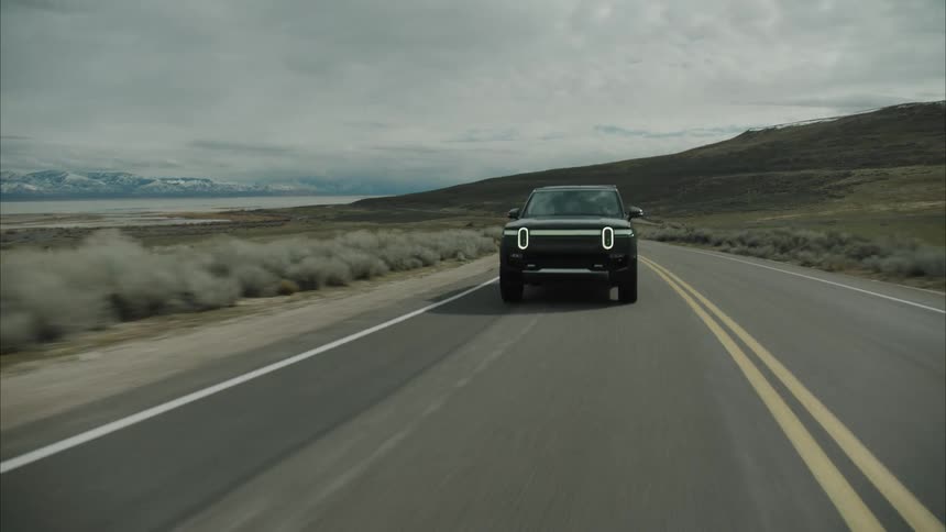 A dark rivian r1s drives on a paved road through a vast barren landscape under an overcast sky