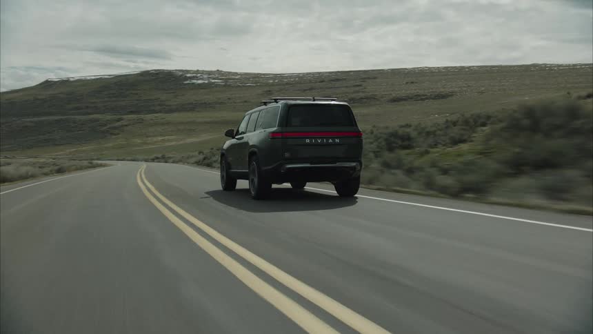 A dark suv drives along a winding asphalt road through a dry hilly landscape under a cloudy sky