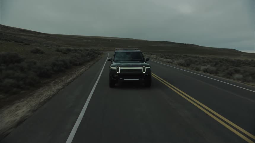 A dark colored rivian r1t drives down a straight asphalt road through a barren hilly landscape under a cloudy sky