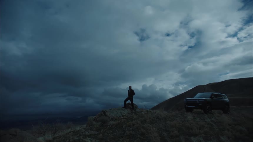 A man steps out of a darkcolored suv parked on a hilltop stands on a rock and gazes at the dramatic landscape under a cloudy sky