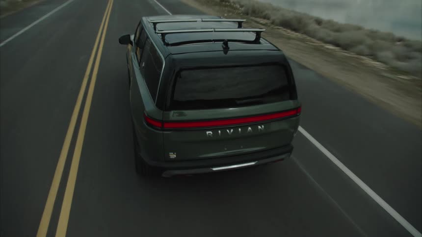 A dark suv drives down a straight asphalt road adjacent to a large body of water with distant mountains visible under an overcast sky