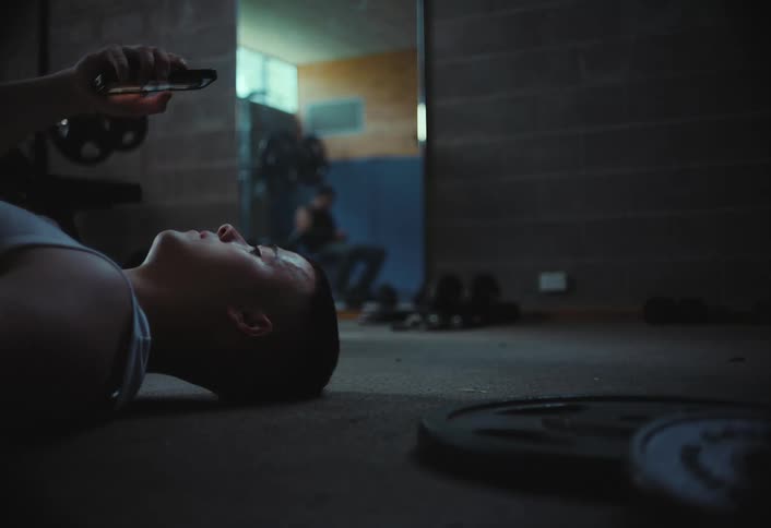 A buzz cut person lies on the gym floor browsing their phone in a dimly lit room