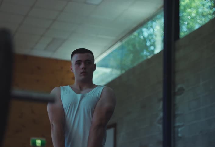 A young man in a white undershirt examines his arm muscles in a gym mirror with sunlight streaming through windows