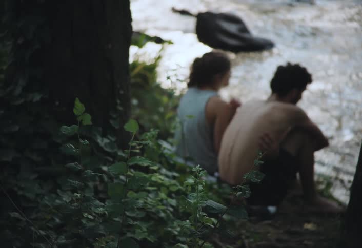 A young couple sits on the riverbank in a forest engaged in conversation