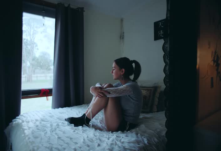 A teenager in shorts sits on a bed with ruffled sheets arms wrapped around their legs gazing out a window in a bedroom appearing deep in thought and lonely