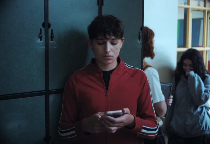 A teenager in a red sweater reclines against school lockers texting on his phone