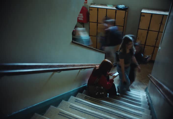 A girl sits on a staircase using her phone while students move around her quickly