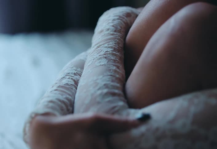 Young woman wraps her arms around her raised knees while sitting on the bed