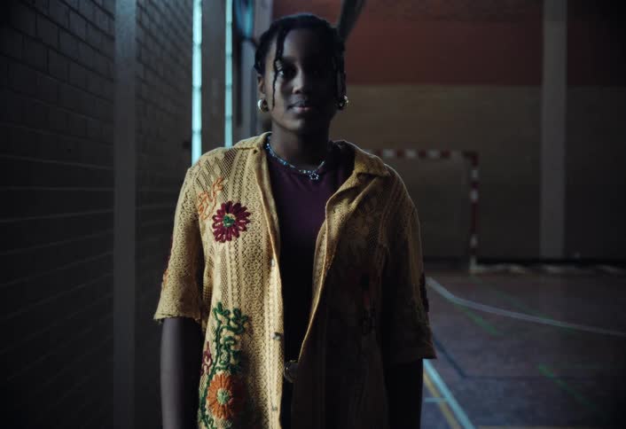 A teenage girl in a yellow cardigan with floral embroidery stands in a dimly lit indoor sports hall
