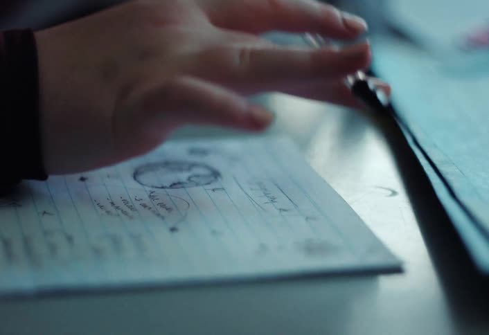 A closeup of a hand balancing a pen over a desk with paper showing handwritten squiggles nearby