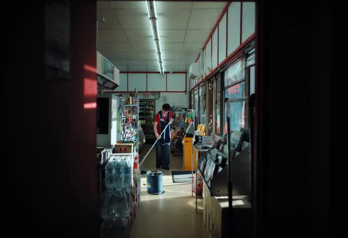 A person is mopping the floor inside a convenience store surrounded by shelves stocked with various products with a mop bucket nearby