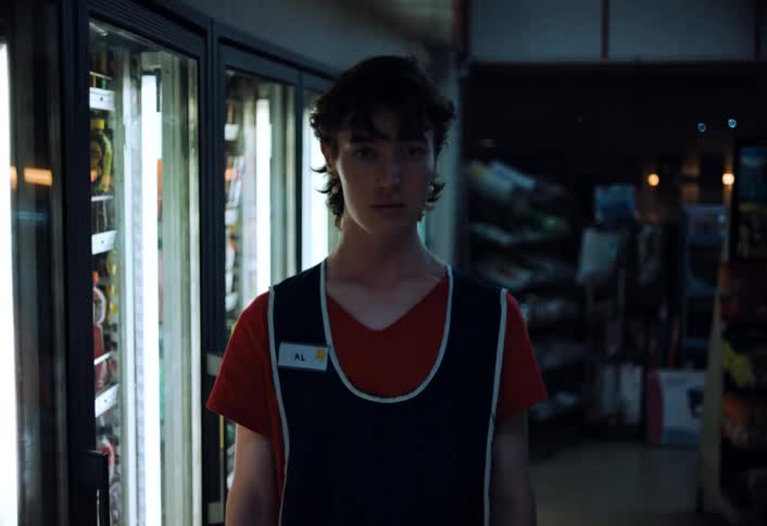 A store clerk stands motionless as the lights go out and return flickering while looking directly at the camera