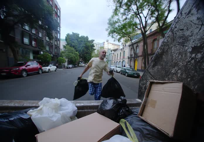 A man in pajamas runs after a trash truck holding bags in his hands as the truck moves away without stopping
