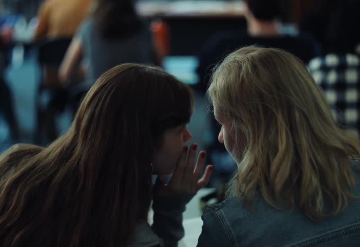 Two girls from behind sitting at a desk whispering something in each others ears