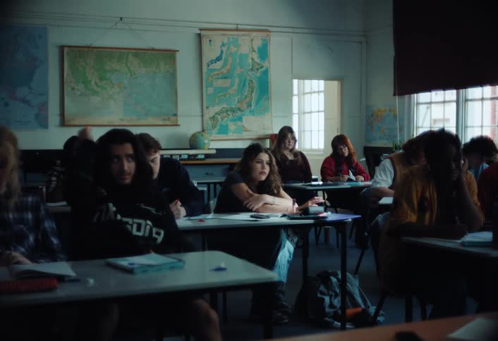A classroom of diverse teenagers sits at their desks some looking engaged while others are lost in thought
