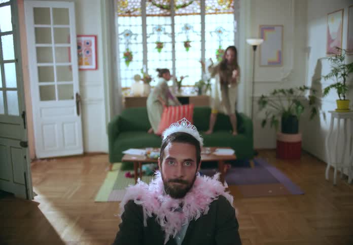 Father playing dress up with his daughters throws a feather boa over his shoulder while his daughters jump on the couch during a pillow fight