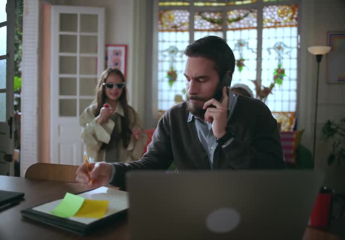 A father is talking on the phone and using his laptop when his daughters come to distract him