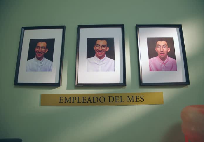 A wall display features three framed employee of the month photos of the same man with one showing him surprised or distressed due to his shirt being pink instead of white