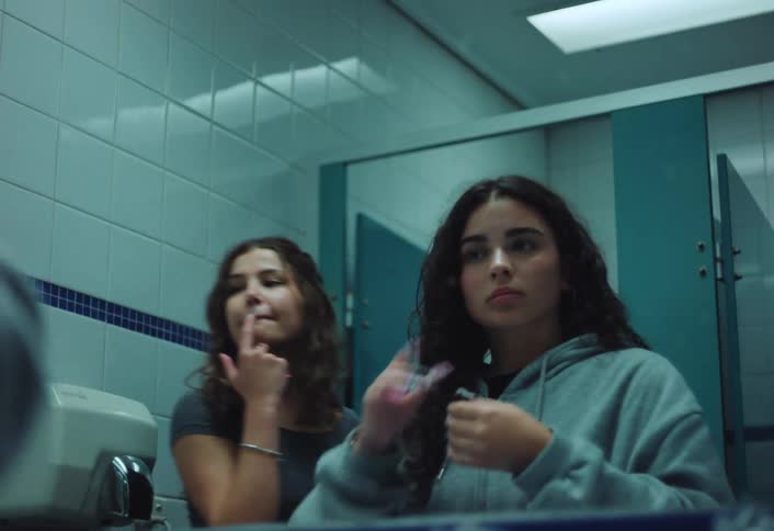 Two teenage girls do their makeup and hair in front of a mirror in a public bathroom